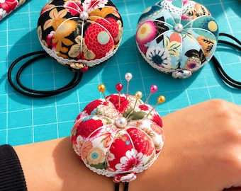 May include: Three round, fabric-covered pincushions featuring floral patterns in black, light blue, and white. Each cushion has a central white bead, white lace trim, and a black elastic wrist strap. One cushion is shown worn on a wrist with pins.