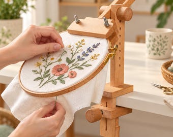 May include: A wooden embroidery hoop stand with a clamp mechanism holding a circular frame. The fabric inside features a hand-stitched floral design with pink, yellow, white, and blue flowers and green leaves on a white textured cloth background.