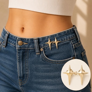 Peut inclure: Une épingle de serrage de taille en métal doré composée de deux étoiles à quatre branches reliées par une charnière centrale. L'accessoire est fixé à la ceinture d'un jean en denim bleu foncé pour ajuster la taille du vêtement.