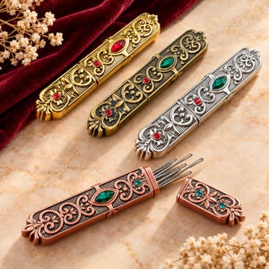 May include: Four rectangular metal needle cases with ornate embossed scrollwork and inset faceted gemstones. Three cases are closed in gold, bronze, and silver tones, while one copper-toned case is open, revealing several thin metal needles inside.