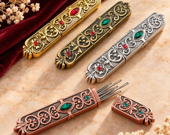 May include: Four rectangular metal needle cases with ornate embossed scrollwork and inset faceted gemstones. Three cases are closed in gold, bronze, and silver tones, while one copper-toned case is open, revealing several thin metal needles inside.