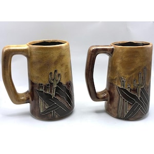 May include: Two ceramic mugs featuring a two-tone glaze with a light tan upper section and a dark brown lower section. The lower portion displays a relief design of a saguaro cactus and desert foliage. Each mug has a large, angular handle.