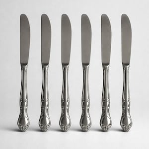 May include: Six stainless steel butter knives with a decorative handle design. The knives have a curved blade and a detailed, ornate handle. The knives are arranged in a row against a white background.