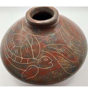 May include: A round, terracotta-colored ceramic vase with a narrow neck. The vase features white etched designs of a turtle and other abstract shapes. The background has red, brown, and blue hues.