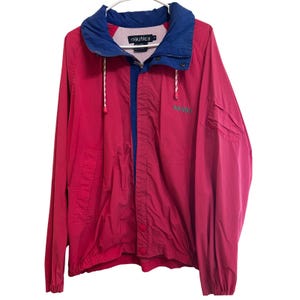 May include: A bright pink windbreaker jacket featuring a contrasting blue collar and inner lining. The jacket includes a front zipper closure, two snap buttons, white drawstring cords, and the Nautica brand name embroidered in green on the chest.