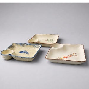 May include: Three ceramic serving dishes with attached sauce bowls. The dishes are square with rounded edges and feature hand-painted designs, including a blue fish, floral patterns, and a wave border. The dishes are off-white with brown trim.
