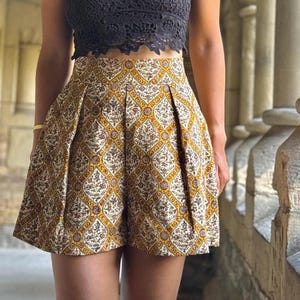 May include: A black lace crop top paired with high-waisted shorts featuring a yellow, brown, and white geometric pattern. The shorts have a pleated design. The outfit is accessorized with sunglasses and a necklace.