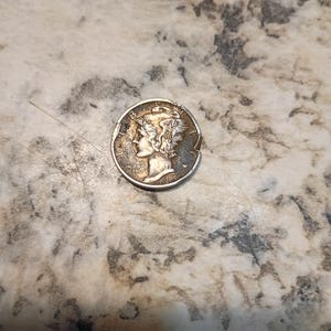 May include: A silver Mercury dime coin, featuring a profile of a woman with a winged cap. The coin is slightly worn, with visible details of the design and the year 1944. The coin is on a speckled, light-colored surface.