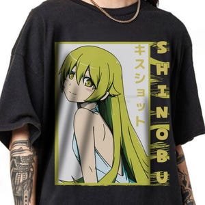 May include: Black t-shirt featuring a colorful anime graphic. The graphic includes a female character with long, light green hair and the Japanese text "キ ス ショ ッ ト" and the word "SHINOBU" in vertical text. The character is wearing a light blue dress.