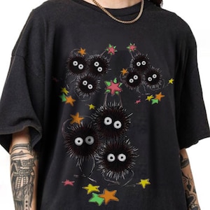 May include: Dark gray t-shirt featuring a whimsical illustration of several black, fluffy creatures with large white eyes and small legs. The design includes colorful stars in shades of orange, pink, and green, creating a playful and unique aesthetic.
