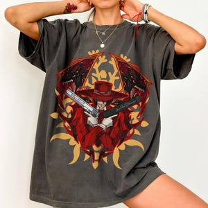 May include: Dark gray t-shirt featuring a graphic of a character in a red suit and hat, holding two pistols. The design includes a red and gold emblem with text on the guns. The shirt is a casual wear item.