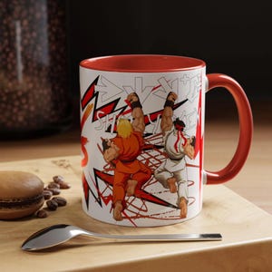 May include: A white ceramic mug with a red interior and handle. The mug features a graphic of two characters in fighting poses, with red and black accents. The mug is on a wooden surface with a spoon and a pastry.