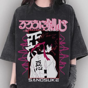 May include: Dark gray t-shirt featuring a Japanese anime-style graphic. The design includes Japanese characters, a stylized character illustration, and the word "SANDSUKE" in white block letters. Pink and white accents.
