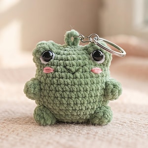 Könnte beinhalten: Ein salbeigrüner, gehäkelter Frosch-Schlüsselanhänger mit großen schwarzen Kunststoffaugen, gestickten rosa Wangen und einem kleinen V-förmigen Mund. Die ausgestopfte Figur hat einen runden Körper, kurze Gliedmaßen und einen silbernen Metallring.