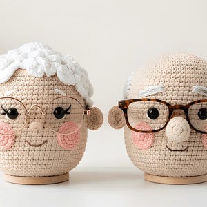 Puede incluir: Se muestran dos muñecos de ganchillo, que se asemejan a una pareja de ancianos. Los muñecos tienen la piel clara, mejillas rosadas y llevan gafas. Uno tiene el pelo blanco y el otro gris. Están colocados sobre bases de madera.