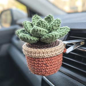 Pode incluir: Uma planta suculenta de crochê num pequeno vaso multicolorido preso a um clipe de metal prateado. A planta apresenta folhas verdes em camadas, enquanto o vaso consiste num rebordo superior castanho escuro, uma faixa bege e uma base de terracota.