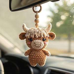 Pode incluir: Um acessório de carro em forma de vaca highland em croché pendurado no espelho retrovisor. A figura é castanho-clara com focinho, franja e chifres cor de creme. Possui olhos pretos, bochechas bordadas a rosa e um pequeno sorriso, suspenso por um anel de madeira e três contas de madeira.