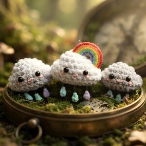 May include: Three small, white, crocheted cloud figures with black bead eyes and pink stitched mouths. Each cloud has dangling pastel-colored raindrops. The center cloud features a small, colorful, semi-circular rainbow attached to its top.