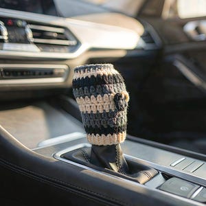 May include: A cylindrical crochet gear shift cover featuring horizontal stripes in cream, dark gray, and black yarn. The cover has a textured, ribbed stitch pattern and includes a small, dark circular crochet accent attached to the side.