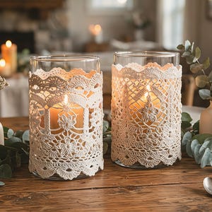 May include: Two cylindrical glass candle holders wrapped in intricate cream-colored crochet lace covers. Each holder contains a lit pillar candle. The set is arranged with small ceramic vases, green eucalyptus sprigs, and white floral accents.
