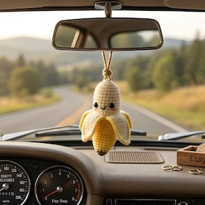 May include: A crocheted banana car charm hangs from a car's rearview mirror. The banana is yellow and cream-colored with a smiling face. The car's dashboard has gauges, a small wooden box with "Crafty Etsy Shop" on it, and gold rings.