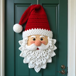 Op de afbeelding: Een handgemaakte gehaakte deurdecoratie van een kerstman. Het gezicht van de kerstman heeft een rode muts met een witte pompon, een witte baard en een roze gezicht. De decoratie hangt aan een teal groene deur.