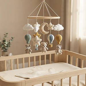 May include: A hanging nursery mobile featuring crocheted elements suspended from a circular wooden frame. The mobile includes white clouds, a crescent moon, smiling stars, hot air balloons in blue and yellow, and three small beige teddy bears.