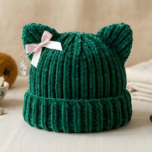 May include: A dark green velvet beanie with cat-ear-shaped points at the top. The hat features a ribbed texture and a small, light pink ribbon bow attached to the left side, accented with a silver-toned charm featuring two small star shapes.