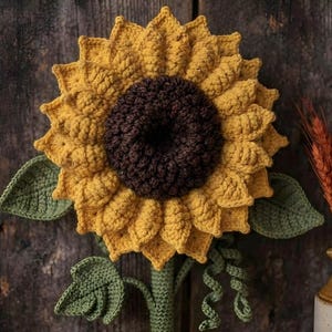 May include: A crocheted sunflower wall hanging featuring a large, textured dark brown center surrounded by vibrant yellow petals, with green crocheted leaves and a stem. The sunflower is displayed alongside a ceramic pitcher containing dried wheat.