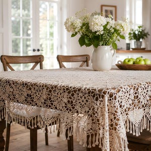 May include: A rectangular cream-colored crochet tablecloth featuring an intricate floral pattern and a decorative fringe border. The textile is draped over a wooden dining table, showcasing detailed needlework and a textured, open-weave design.