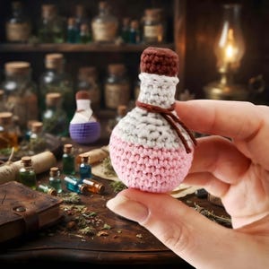 May include: A small, handcrafted crochet potion bottle featuring a rounded base in light pink, a white middle section, and a dark brown cap. A thin brown yarn string is tied around the neck of the bottle, which is held against a blurred background.