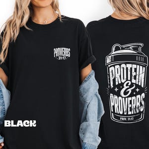 May include: A black t-shirt featuring a white graphic on the front left chest that reads "PROVERBS 31:17". The back of the shirt displays a large white graphic of a protein shaker bottle with the text "PROTEIN & PROVERBS PROV. 31:17" in a distressed font.