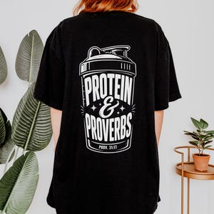 Puede incluir: Camiseta negra con un gráfico blanco de un mezclador de proteínas y las palabras "PROTEIN & PROVERBS". El texto "PROV. 31:17" está debajo. La camiseta es de estilo casual.