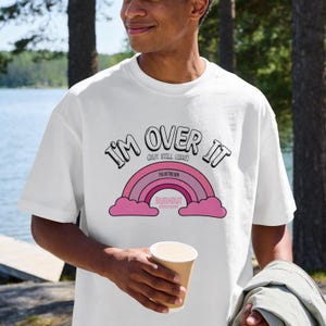 May include: White t-shirt with the text "I'M OVER IT (BUT STILL HERE)" above a pink rainbow design. The shirt also features the text "THE RETRO CREW" and "BURNOUT EDITION".