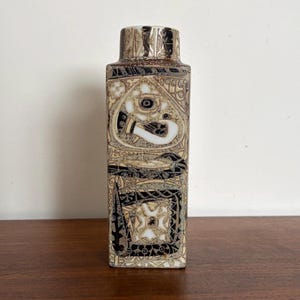 May include: A rectangular ceramic vase featuring a complex, intricate pattern in shades of cream, beige, and black. The surface is covered in detailed geometric and abstract motifs, including stylized eye shapes and ornate borders, with a narrow neck.