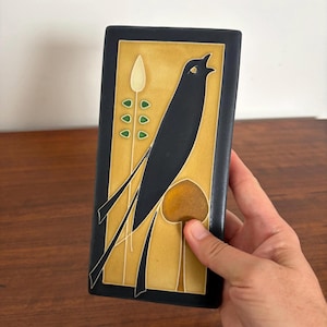 Puede incluir: Un azulejo de cerámica rectangular que presenta la silueta de un pájaro negro sobre un fondo amarillo mostaza. El diseño incluye una planta alta y delgada con pequeños detalles de hojas verdes y una forma marrón redondeada, bordeado en negro.