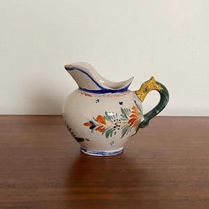 Pode incluir: Jarro de cerâmica pequeno, creme, com corpo arredondado e bico. Decorado com padrões florais laranja, verdes e azuis. A pega é verde com um detalhe dourado. O jarro está sobre uma superfície de madeira.