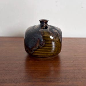 May include: A small, squat ceramic vase with a narrow neck and a rounded body. The surface features a glossy, olive-green glaze with horizontal ridges on the lower half and a dark, textured, matte-black glaze covering the upper portion.