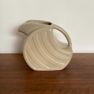 May include: A beige ceramic pitcher with a curved handle and spout. The pitcher has a rounded body with a series of parallel, textured lines. The pitcher is sitting on a dark wooden surface.