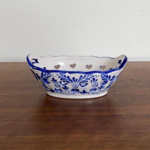 Może przedstawiać: Mała, biała ceramiczna miska z niebieskimi wzorami kwiatowymi i wycięciami w kształcie serca. Miska ma falisty brzeg i dwa małe uchwyty. Niebiesko-biały wzór przypomina ceramikę z Delft.