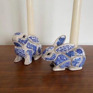 May include: Two ceramic bunny candle holders with blue and white floral and geometric patterns. Each bunny holds a cream-colored candle. The candle holders are on a dark wooden surface.