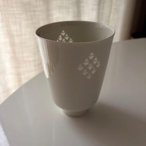 以下が含まれることがあります： 白いセラミックカップ。上部はフレア、下部は小さなベース。表面には小さな三角形の幾何学模様が施されています。白い表面に置かれたカップ。