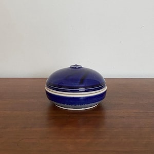 Könnte beinhalten: Kleine, runde Schmuckdose aus Keramik mit Deckel. Die Dose ist tiefblau mit einem weißen Streifen in der Mitte. Der Deckel hat einen kleinen Knopf oben. Die Dose steht auf einer Holzoberfläche.