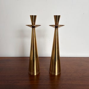 May include: A pair of tall, polished brass candlesticks featuring a slender, conical base that tapers upward to a wide, flat circular drip plate and a narrow candle cup. The metal surface has a reflective gold-toned finish with fine horizontal lines.