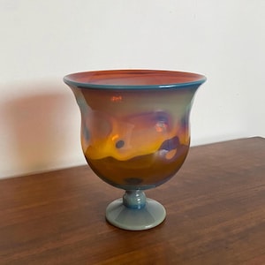 Può includere: Un vaso in vetro iridescente fatto a mano, con una parte superiore ampia e svasata e una base a piedistallo. Il vaso presenta una miscela di colori, tra cui arancione, giallo e blu, creando un aspetto artistico unico. È un oggetto decorativo.