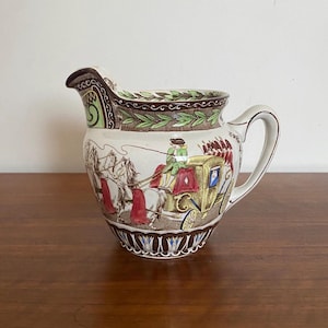 Op de afbeelding: Crèmekleurige keramische kan met een handvat en tuit. De kan heeft een gedetailleerde illustratie van een paardenkoets met figuren in historische kleding. De rand is versierd met groene bladeren en bruine accenten. Een klassiek, vintage decoratief stuk.