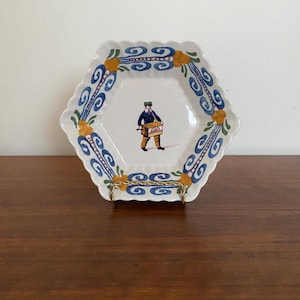 May include: A white, hexagonal decorative plate with a scalloped edge. The plate features a blue and yellow scroll design around the rim and a central illustration of a person in a blue jacket and brown trousers.