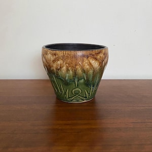 Puede incluir: Una maceta de cerámica con interior oscuro y exterior texturizado. La maceta presenta un degradado de color, que va del marrón en la parte superior al verde en la inferior. El diseño incluye un patrón floral o de hojas estilizado.