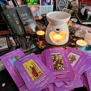 Puede incluir: Una colección de cartas del tarot con dorsos morados e ilustraciones, junto con un difusor de aceites esenciales blanco con recortes en forma de corazón. También hay velas encendidas y cristales, creando una atmósfera mística.