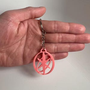 May include: A light pink keychain with a circular design and a starburst-like pattern inside. The keychain is attached to a silver chain and ring. The design is a simple, geometric shape.